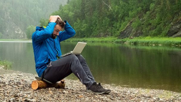 A guy in a blue jacket with a white laptop is sitting on a log in the rain on the bank of the river and holding his head. Conceptual error in programming or disruption of a business transaction. - Foto, afbeelding