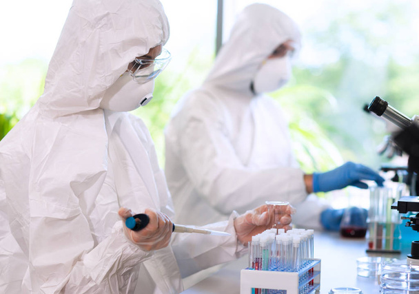 Scientists in protection suits and masks working in research lab using laboratory equipment: microscopes, test tubes. Biological hazard, pharmaceutical discovery, bacteriology and virology concept. - 写真・画像