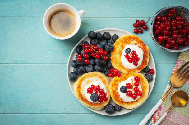 Cottage cheese pancakes with red currant and blueberry. Coffee, berries and sauce on blue background. Fresh and tasty breakfast. - Foto, Imagen