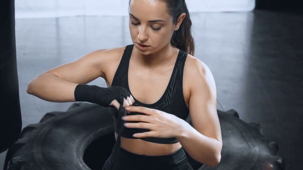 beautiful sportswoman sitting on tire and putting on training gloves in gym - Footage, Video