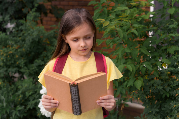 portrait of a little beautiful girl with a backpack holding a book and reading  on the background of a brick wall and green bushes - Φωτογραφία, εικόνα