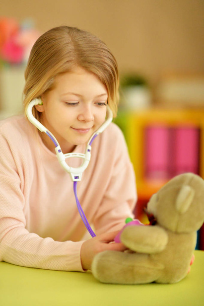 Cute little girl playing nurse, inspecting teddy bear with stethoscope - Photo, Image