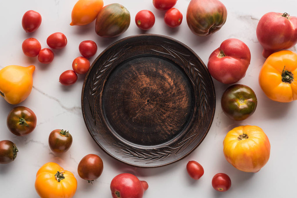 top view of empty wooden plate near scattered tomatoes on white marble surface  - Photo, Image