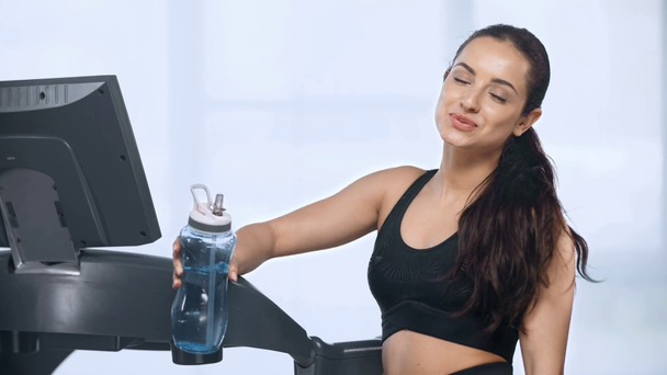 beautiful sportswoman near treadmill looking at camera, smiling and drinking water in gym - Video