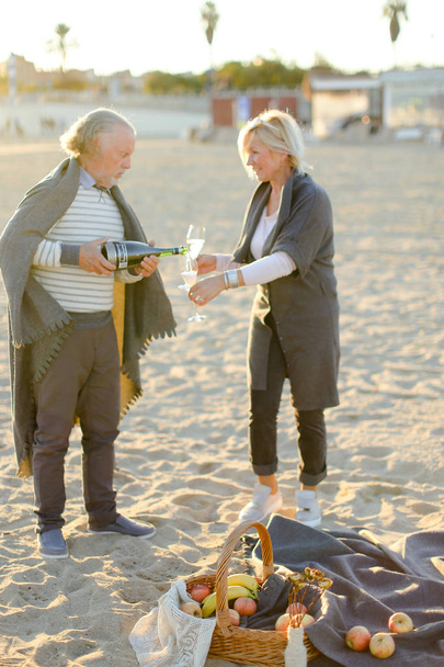 Focus on fruits and plaid, senior pouring champagne with wife on sand beach in background. - Photo, Image