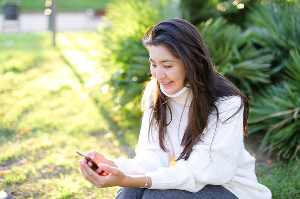Κινέζικο κορίτσι κάθεται στο πάρκο και χρησιμοποιώντας smartphone. - Φωτογραφία, εικόνα
