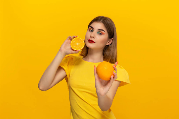 Young model in a yellow tee holds oranges standing over yellow background. - Фото, изображение