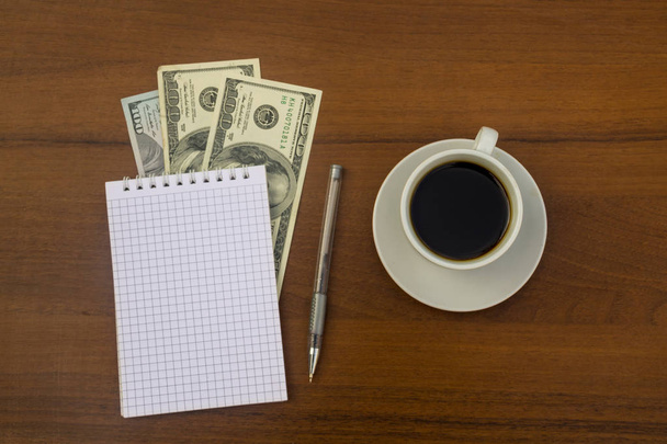 Cup of coffee, dollars, notepad and pen on wooden desk - Photo, Image
