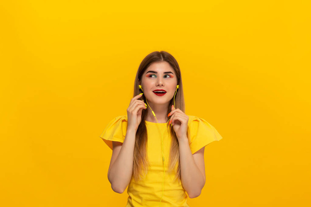 Happy young model in a yellow tee listens to music with wired headphones standing over yellow background - Photo, Image