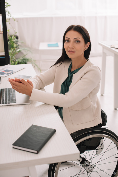 beautiful disabled businesswoman smiling at camera while sitting at workplace in wheelchair - Φωτογραφία, εικόνα