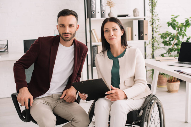 positive disabled businesswoman holding clipboard while sitting near business partner in office - Foto, Imagem