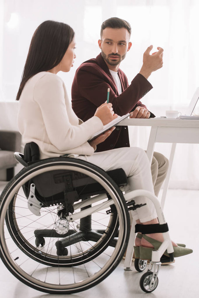 empresaria discapacitada sosteniendo lápiz y cuaderno mientras habla con su socio de negocios en la oficina
 - Foto, imagen