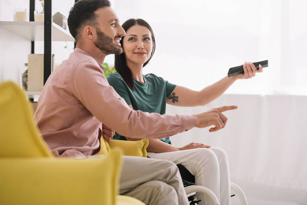 cheerful man pointing with finger while watching tv together with disabled girlfriend - Foto, Bild
