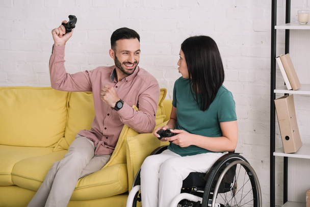 KYIV, UKRAINE - JULY 10, 2019: Happy young man showing winner gesture while playing video game with disabled girlfriend. - Φωτογραφία, εικόνα