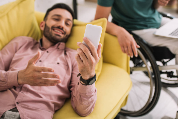 ラップトップを使用して障害のあるガールフレンドの近くにスマートフォンでソファに横たわっている陽気な男 - 写真・画像