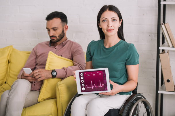 hübsche, behinderte Frau zeigt digitales Tablet mit Herzfrequenz-App, während sie neben Freund mit Smartphone sitzt - Foto, Bild