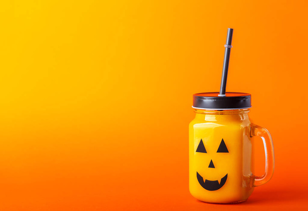 Halloween healthy pumpkin or carrot drink in the glass jar with scary face on a orange background with copy space - Фото, изображение