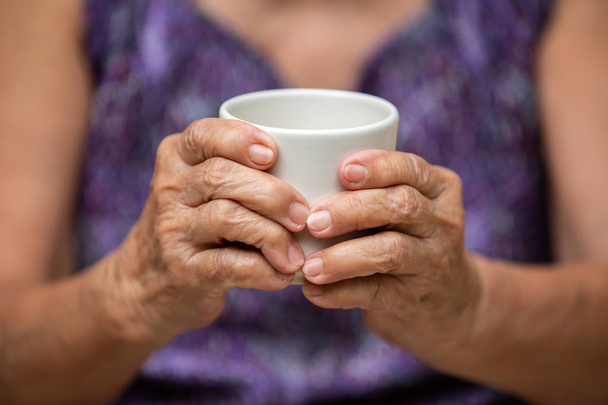 Mani della donna anziana che tengono in mano tazza di tè aromatico in bokeh sfondo vestiti viola, parte asiatica della pelle del corpo, Relax time & Drink concept
 - Foto, immagini
