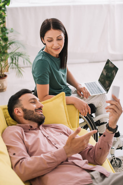 sonriendo mujer discapacitada usando el ordenador portátil mientras está sentado cerca de novio acostado en el sofá con teléfono inteligente
 - Foto, Imagen