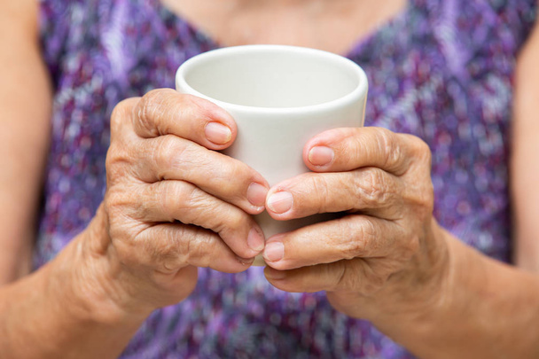 Las manos de la mujer mayor sosteniendo la taza de té aromático en el fondo de la ropa de color púrpura bokeh, parte de la piel del cuerpo asiático, tiempo de relajación & concepto de bebida
 - Foto, Imagen