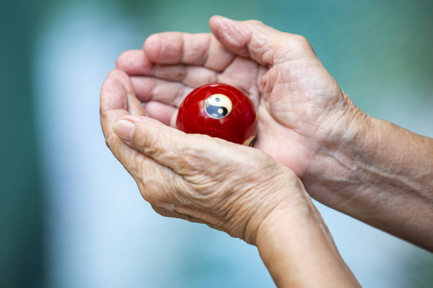 Senior woman's hands holding and praying a black and white Yin Yang religious symbol on red ball in Taoism in bokeh blue swimming pool background, Asian Body skin part, Body language feeling, Religion concept - Photo, Image