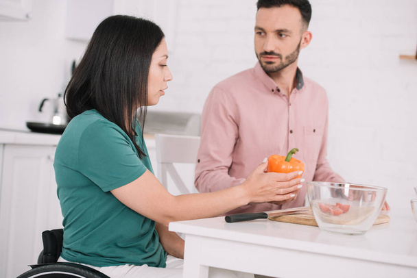 junge behinderte Frau hält Paprika in der Hand, während sie mit Freund in der Küche spricht - Foto, Bild