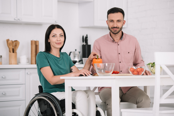 atractiva mujer discapacitada con novio sonriendo a la cámara mientras se sienta en la mesa de la cocina y preparar ensalada juntos
 - Foto, Imagen