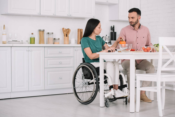 joven mujer discapacitada con novio guapo sentado en la mesa de la cocina y preparar ensalada juntos
 - Foto, Imagen