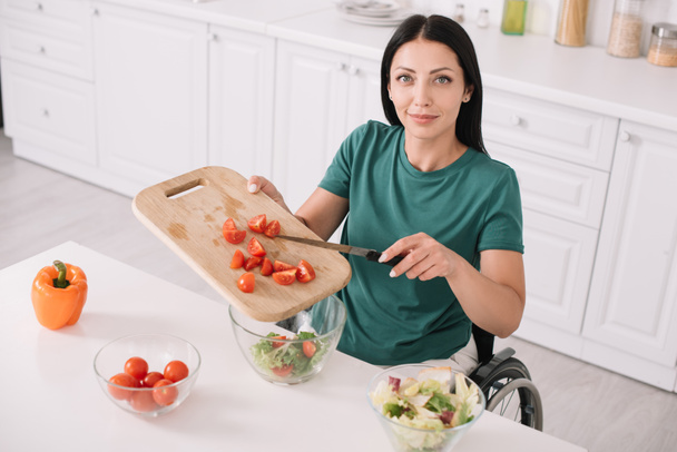 cheerful disabled woman looking at camera while adding sliced tomatoes in bowl - Zdjęcie, obraz