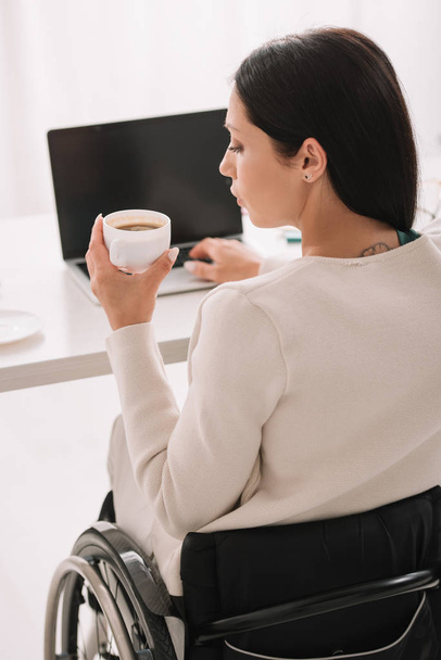 vista posterior de la mujer de negocios discapacitada sosteniendo la taza de café mientras usa el ordenador portátil en la oficina
 - Foto, imagen