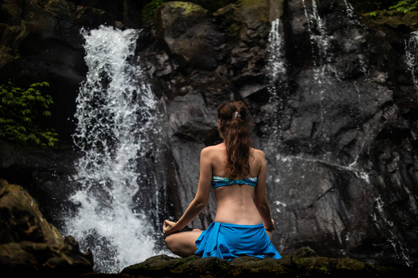 Nuori kaukasialainen nainen meditoimassa, joogaamassa vesiputouksella Balilla, Indonesiassa. Näkymä takaa
. - Valokuva, kuva