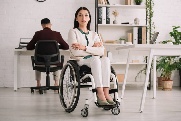 zelfverzekerde gehandicapte zakenvrouw zittend in rolstoel met gekruiste armen in de buurt van collega werken op achtergrond - Foto, afbeelding