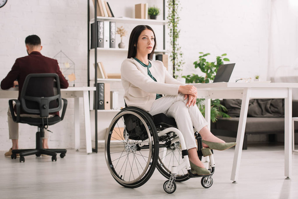 serious disabled businesswoman sitting in wheelchair in office near colleague  - Foto, Imagen