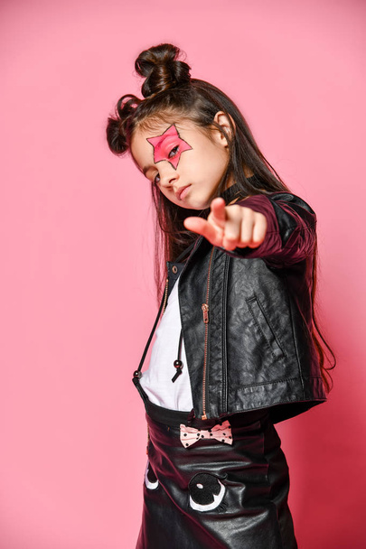 fashionable hipster punk girl - dressed in a leather jacket and skirt, black knee-highs and a white T-shirt, with a funny hairstyle and a makeup painted star on her face, points to you. - Фото, изображение