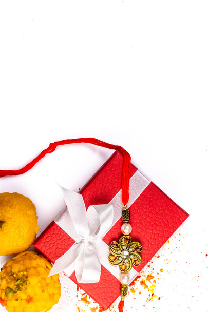 Индийский фестиваль - Top view of elegant Rakhi on a red gift box with sprinkled rice, kumkum and haldi and sweets on white background
 - Фото, изображение