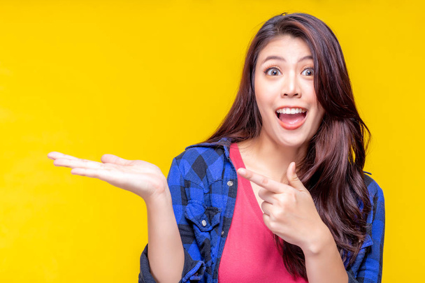 Beautiful young asian woman pointing finger to copy space. Attractive beautiful girl show product that make her satisfied, excited, love, surprise with exciting face, surprise face. Yellow background - Photo, Image