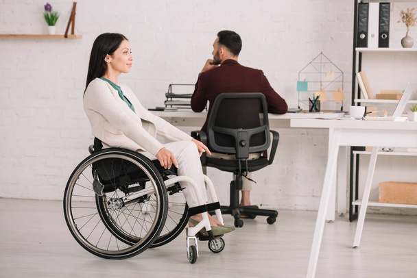smiling disabled businesswoman looking away while sitting in wheelchair near colleague - Foto, Imagen