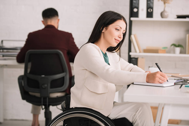 cheerful disabled businesswoman writing in notebook while sitting in wheelchair at workplace - Zdjęcie, obraz