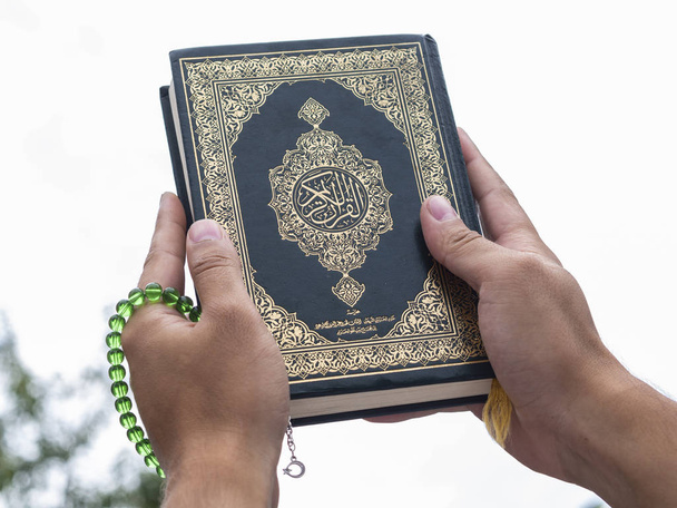Mans handen houdt Koran-heilig boek van moslims met islamitische rozenkrans, op blauwe hemel met wolken - Foto, afbeelding