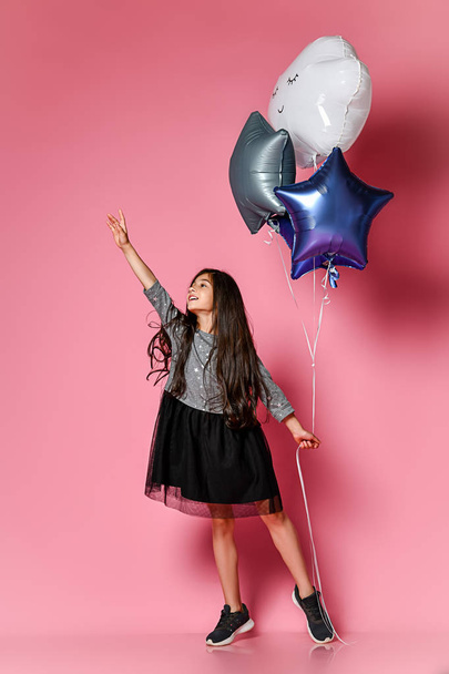 beautiful young girl with long dark hair in a dress holds pink balloons in the shape of stars and clouds and is looking at the camera against a pink background - Photo, Image