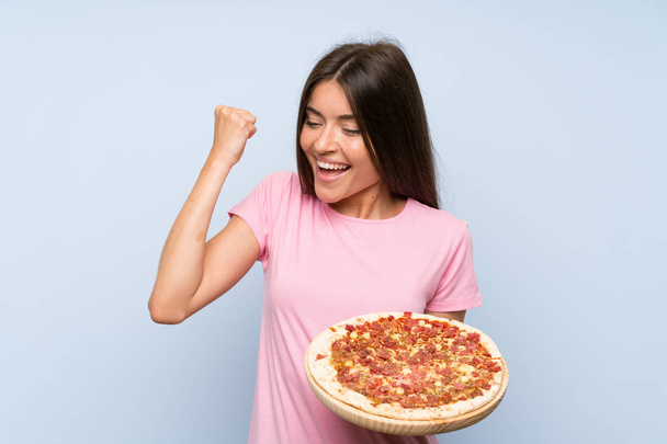 hübsches junges Mädchen, das eine Pizza über eine isolierte blaue Wand hält und einen Sieg feiert - Foto, Bild