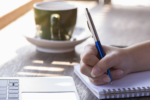 Hand hält einen Stift in der Nähe eines Notizbuchs mit einem Kaffeebecher daneben auf einem hölzernen - Foto, Bild