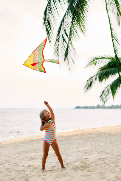 Happy little girl with flying kite on tropical beach under palm trees. Beach fun, happy childhood concept                   - Valokuva, kuva