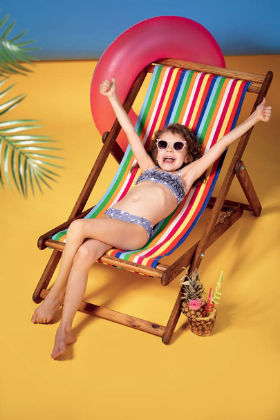 Dziewczyna ubrana w okulary przeciwsłoneczne i strój kąpielowy opalając się w tęczowym krześle. Podniesione ręce i uśmiech - Zdjęcie, obraz
