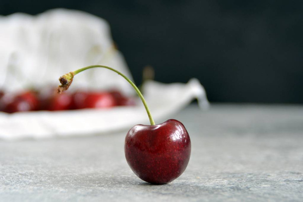 A single cherry lies on a gray marble surface and is focussed, in the background are many cherries in a bowl  - Photo, Image