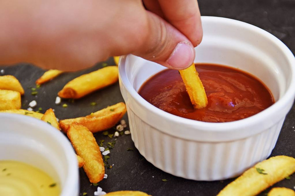  fresh potato fries of golden yellow fried in a bowl full of ketchup, against a dark marble background, on which fries and salt grains and spices lie - Valokuva, kuva