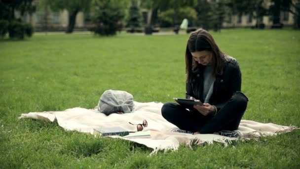 Charming female student learning outdoors at campus using a tablet. Student sitting with cross legged on blanket on green grass, video - Footage, Video