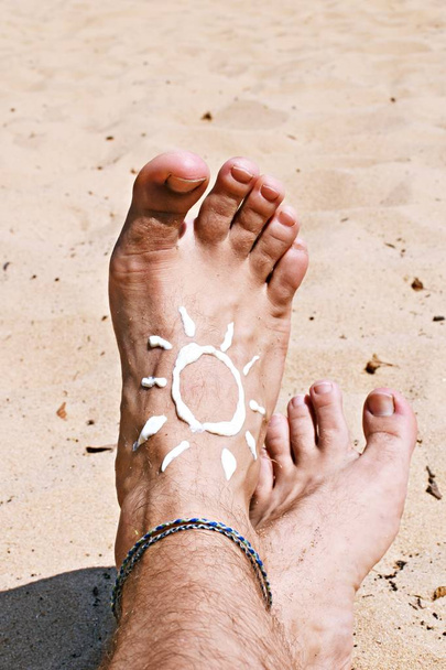 На босой ноге с солнцезащитным кремом, солнце было нарисовано на фоне песка с пляжа, чтобы увидеть - Концепция защиты от солнца в жаркие дни пляжа с мужской ногой с браслетом
 - Фото, изображение