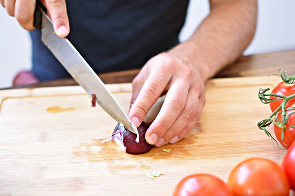 Ένα άτομο κόβει ένα κόκκινο κρεμμύδι σε μια ξύλινη σανίδα για μια φρέσκια τραγανή υγιεινή σαλάτα-κοντά στα χέρια του ατόμου-έννοια για την υγιεινή διατροφή με φρέσκα βιολογικά λαχανικά - Φωτογραφία, εικόνα