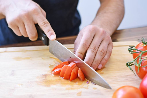 A person cuts a tomato on a wooden board for a fresh crunchy healthy salad - close-up on the hands of the person - concept for healthy nutrition with fresh organic vegetables  - Fotoğraf, Görsel